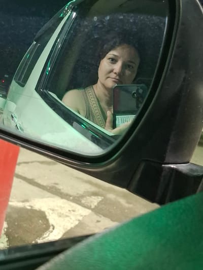 A medium shot photograph captures a woman taking a selfie in a car's side mirror. The woman, with short brown hair and a neutral expression, is looking directly into the camera. She is wearing a light-colored tank top with a patterned strap across her shoulder. Her left hand is visible, holding a dark-colored smartphone with multiple camera lenses. The background in the mirror shows the interior of the car, with a slightly blurred view of the driver's side window and door. The car appears to be parked, as indicated by the surrounding environment. The exterior of the car is reflected in the mirror, showing a portion of the vehicle's body. The foreground of the image is dominated by the car's side mirror casing and a part of the car's body, painted in dark and light colors, respectively. A glimpse of the exterior environment is visible in the reflection on the car's body and in the lower part of the image, which appears to be a road or parking lot, possibly with wet patches. The image is taken at night or in low light conditions, as suggested by the darkness of the background and the reflections. The location is identified as Svobodny, Russia.
