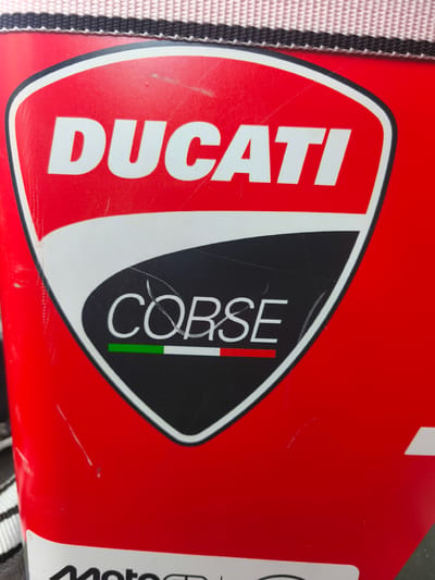 The image shows a close-up of a red object, likely a bag or piece of equipment, featuring the DUCATI CORSE logo. The logo is set against a red background with white and black accents. The DUCATI text is prominent in white lettering. Below it, the CORSE text is also in white. Underneath the CORSE text, there's a stripe of green, white, and red, representing the Italian flag. The object has visible scratches and scuffs, indicating wear and tear. The top edge of the object reveals a striped pattern in black and white. There is no visible indication of the city of Nàquera / Náquera, Spain within the image itself, nor any people or specific actions taking place.