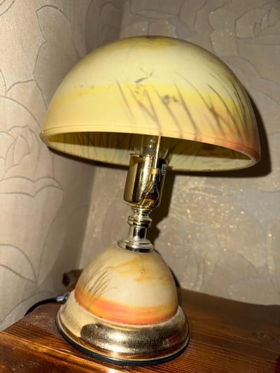 An ornate, vintage-style table lamp with a dome-shaped shade sits on a wooden surface. The shade is adorned with a gradient of yellow and orange, with stylized black lines resembling tall grass or reeds. The lamp's base has a similar design and a metallic, golden finish. The background appears to be wallpaper with a floral pattern. The scene is lit from the lamp itself, casting a warm glow and shadows. The image does not provide any information about the city of Shamkhal, Russia, or any people, actions, or interactions within the scene.