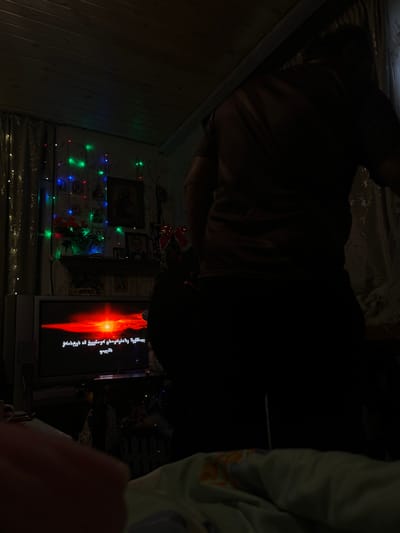 An interior scene is depicted in a dimly lit residential room in Lanchkhuti, Georgia. In the immediate foreground, a blurred human finger is visible in the bottom-left corner, suggesting a handheld camera perspective. The bottom of a light-colored, patterned blanket or bedding occupies the extreme lower portion of the frame.
A silhouette of an individual, seen from behind and facing left, occupies the central and right foreground. The person wears a dark, short-sleeved top and dark lower garments; specific features are obscured by shadow.
To the left, a television screen displays a vibrant red and orange sunset or sunrise over a dark landscape, with white Georgian text overlaying the image. The text reads "შორს სადმე მივდიოდი შორს სულ სხვაგან", which translates to "I was going somewhere far away, somewhere completely different." Below the television, a dark, mesh-like object is partially visible.
Above the television and extending along the wall, multi-colored string lights (red, green, blue) are draped, illuminating a shelf unit. On this shelf, several framed objects are visible, including what appears to be a religious icon with a gold-colored halo and at least two other framed pictures. A vase containing artificial red and orange flowers is also on the shelf. To the right of the shelf, a large red decorative bow is present. Additional white or warm-toned string lights are visible on light-colored curtains along the far-left wall. The ceiling features light-colored wooden paneling. Sheer, patterned curtains are also visible on the right side of the room, behind the standing individual.