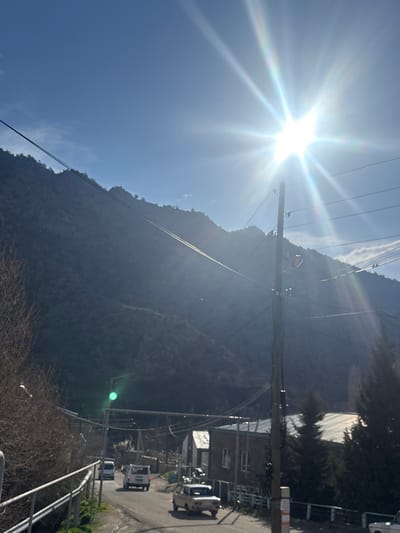 The image depicts a rural or semi-urban road scene in Lehvaz, Armenia, under bright sunlight. A large, dark mountain range dominates the background, rising from the left and center, partially obscured by haze and strong backlighting from the sun. The sky is clear blue with minimal clouds.
In the foreground, a paved or dirt road curves from the lower left, ascending into the mid-ground. Along this road, three light-colored vehicles are visible: two white vans or SUVs further up the road, and a white sedan (possibly an older model) closer to the foreground, facing away from the camera. A metal guardrail or fence lines the left side of the road in the immediate foreground, bordered by bare deciduous trees or bushes.
On the right side of the road, several low-rise buildings with light-colored roofs are present, appearing residential or small commercial structures. Evergreen trees (conifers) are clustered near these buildings. A prominent utility pole, likely wood or concrete, stands in the right-center, supporting multiple overhead power lines or communication cables that traverse the frame horizontally and diagonally.
The sun is a brilliant starburst originating from the upper right quadrant, casting intense light and causing significant lens flare, which creates a bright glow and a green artifact in the lower-central part of the image. The strong sunlight backlights much of the scene, particularly the mountains and portions of the road. No individuals are discernible in the scene.