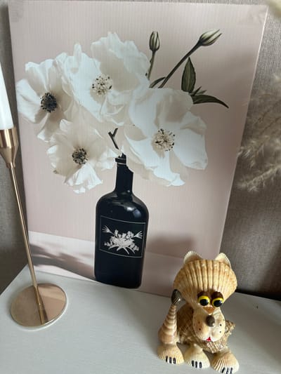 The image displays a still life arrangement on a white surface, likely a shelf or table. The focal point is a canvas print featuring a bouquet of white flowers in a black bottle against a pale pink background. To the left of the canvas, a slender gold candlestick with a white candle is partially visible. In the foreground, to the right of the canvas, sits a whimsical figurine crafted from seashells, resembling an animal with large googly eyes. The background consists of a textured grey fabric. The overall scene is indoors and does not contain any identifiable elements linking it to the city of Рубцово, Russia.