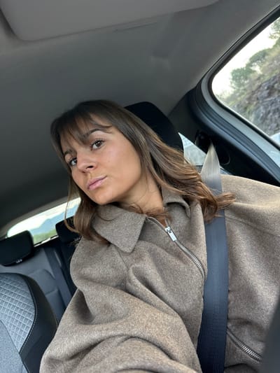 A woman with brown hair and bangs sits in a car, wearing a brown coat with a silver zipper. A dark seatbelt crosses her chest. The interior of the car is visible, including a gray dashboard and seat. Outside the window, a glimpse of trees and sky can be seen. The image is taken at an angle, slightly tilted to the side.