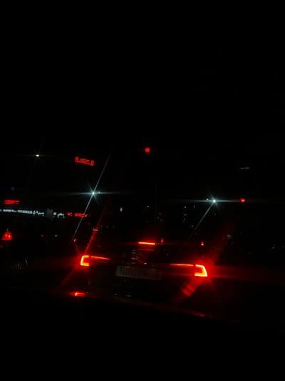 The scene depicts a nighttime street view in Beni Mellal, Morocco, captured from the perspective of a vehicle. The foreground is occupied by the rear of a dark-colored vehicle displaying illuminated red taillights and a legible Moroccan license plate: "74055 | 6". To the immediate left, a building facade presents illuminated signage, including "YSFS MEDICALES" and Arabic text "صيدلية" (pharmacy) alongside a green cross symbol, identifying a medical or pharmaceutical establishment. Above this, another sign reads "L'OCCASION VOYAGES ET LOISIRS." Multiple red vehicle taillights extend into the distance, indicating vehicular traffic. Prominent lens flares from bright artificial light sources, likely streetlights or oncoming headlights, are visible across the frame. A partially obscured red traffic signal is present in the upper right quadrant. The environment is dark, with illumination primarily provided by these artificial light sources. No individuals are clearly discernible.