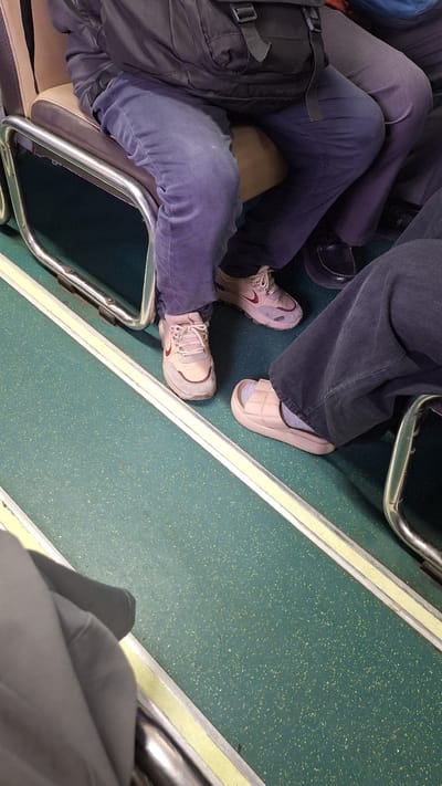 The image captures a close-up perspective of individuals' legs and feet on a green speckled floor, likely within a public transportation setting. Two individuals are visible, one wearing blue pants and light beige athletic shoes with red accents, and another wearing dark pants and light beige slides over grey socks. The floor features a yellow and silver striped border. A portion of a dark bag is visible in the upper left corner, and the seats appear to be upholstered in beige and brown. The lighting suggests an indoor environment, possibly during the day. The scene is static and focuses on the lower bodies of the individuals.