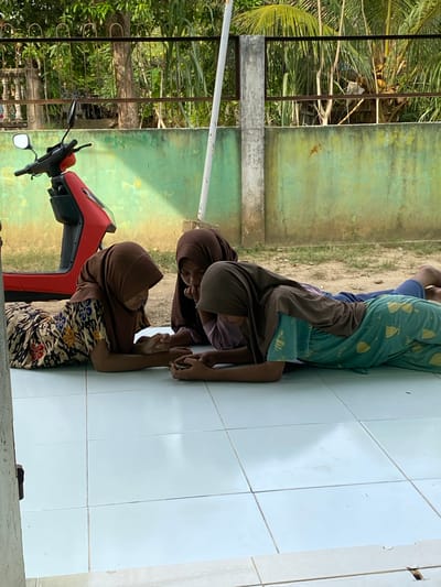 The image, taken in Gampongaceh, Indonesia, shows three individuals resting or observing on a light-colored tiled floor. In the foreground, a young woman or girl lies prone on the tiles, her head resting on her folded arms. She wears a patterned floral dress and a brown hijab. Behind her, another girl in a brown hijab and plain brown top adopts a similar prone position, with her head resting on her arms. Further to the right, a third person, possibly male, is partially visible in a similar posture, wearing a blue shirt and patterned green pants on which the word "Hello" is distinctly legible.

To the left of the foreground individual is the front portion of a red scooter or motorcycle, including its handlebars and mirror. The setting appears to be an outdoor or semi-outdoor area, brightly lit by natural daylight, suggesting it is daytime. Behind the individuals and the scooter, a weathered green wall with some discolored patches forms the backdrop. Beyond this wall, a dark metal fence or railing is visible, leading to a lush tropical environment filled with palm trees and other dense green foliage. The scene conveys a sense of calm and relaxed activity within a warm, natural environment.