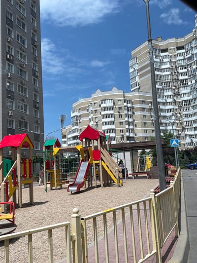 This media file depicts a residential area with modern apartment buildings and a children's playground. The scene is bathed in daylight, suggesting it is daytime, and the bright blue sky with scattered clouds indicates fair weather.
The primary subjects are the towering apartment buildings, characterized by numerous windows and balconies, common in urban Russian architecture. One building on the left has a grey facade with white window frames, while another on the far left features a cream and beige patterned facade. Several air conditioning units are visible on the buildings' exteriors.
In the foreground and mid-ground, a colorful children's playground is set up with various play structures, including slides, swings, and climbing frames, made of wood and brightly colored metal in red, yellow, and green. The ground surface of the playground is covered with fine gravel.
A yellow metal fence with vertical bars lines the right side of the image, separating the playground area from a paved walkway and a street beyond. A blue road sign with a white arrow is visible on a pole near the buildings. A dark blue car is parked on the street in the background.
Although the image is a still photo, the presence of the playground implies that children might be playing there, or have recently done so. The scene evokes a sense of everyday life in a residential urban environment. No people are clearly visible or identifiable as actively engaged in an activity. The location is identified as Azov, Russia. There is no visible text in the image.