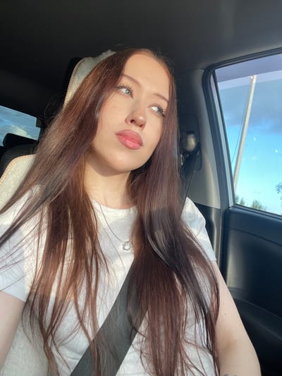 A young Caucasian woman with long, dark brown hair is seated in the passenger seat of a vehicle, looking out the window. She is wearing a white t-shirt and a silver necklace with a heart-shaped pendant. The interior of the car is visible, including the seatbelt, car door, and a portion of the dashboard. Sunlight streams in from the right side of the frame, illuminating her face and hair. The window shows a view of the sky with scattered clouds and some distant trees, suggesting an outdoor location, possibly on the outskirts of a city like Gavrilovka, Russia.