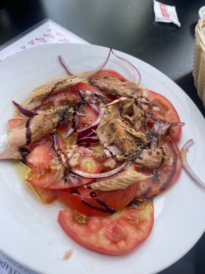 The image depicts a plate of food, specifically a salad with sliced tomatoes, red onion, and what appears to be canned tuna, drizzled with balsamic glaze. The plate is white and sits on a black table. A basket is visible in the upper left corner. A packet of sugar is also visible to the left of the plate. A menu can be seen on the lower right side. The food is centrally located in the frame and illuminated by ambient light. The setting appears to be an outdoor dining area in Jerez de la Frontera, Spain, based on the menu visible.