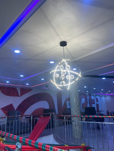 An indoor space featuring a modern, spherical chandelier with illuminated globes. The ceiling is white with blue neon strips and embedded spotlights. A mural adorns the wall, and metal stairs with a red garland and a red fabric drape are visible. Tables and chairs are present in the background.
