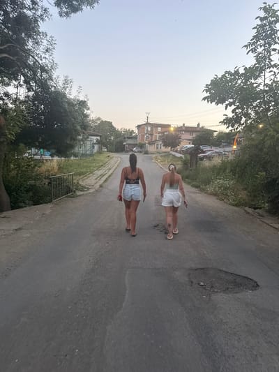In the city of Varvara, Bulgaria, two women walk away from the camera down a narrow, cracked asphalt road. The woman on the left wears a black tank top and denim shorts, carrying a red object in her right hand. The woman on the right wears a light green tank top and white shorts, her left arm extended slightly, holding a dark object. They walk past a metal fence and overgrown vegetation on the left side of the road, and on the right, more vegetation, parked cars, and buildings are visible in the background. The sky is a pale gradient of light blue and white, suggesting either dawn or dusk.