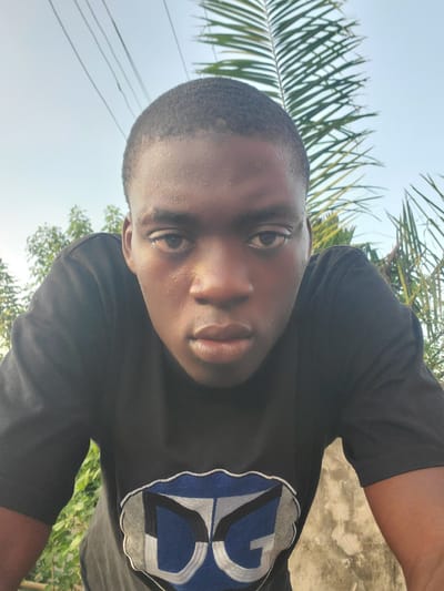 A young African man is taking a selfie outdoors, likely in Atani, Nigeria. He wears a black t-shirt with a blue and white design. The background includes a mix of greenery, palm fronds, and a pale sky. The perspective is from a low angle looking up at his face.