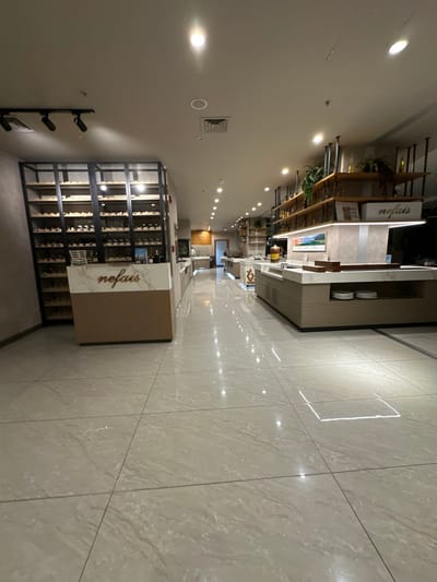 The image displays the interior of a commercial food service or dining establishment in Zeytinburnu, Turkey. The space features light-toned (beige/grey) reflective tiled flooring and a white ceiling equipped with recessed spotlights and black track lighting fixtures.

On the left, a tall, multi-level display unit with a black metal frame and light wooden shelves contains numerous horizontally arranged items, appearing to be bottles or packaged goods. In front of this unit is a two-toned counter, brown/beige on the lower section and white marble with brown veining on the upper surface, bearing the stylized word "nefais." A white basket rests on the counter.

Along the right side, extending into the background, are multiple light-toned buffet or serving stations with light countertops. The station in the foreground includes white plates stacked on a lower shelf. Further back, one station features an illuminated display with a landscape image. Above these stations, open shelving units with black metal frames and wooden shelves display potted greenery, bottles, and decorative boxes, one of which is branded "nefais." A beverage dispenser with a red liquid is present on one of the stations.

One individual is partially visible in the far background, positioned near a doorway, facing away from the camera. No other persons or specific actions are discernible.