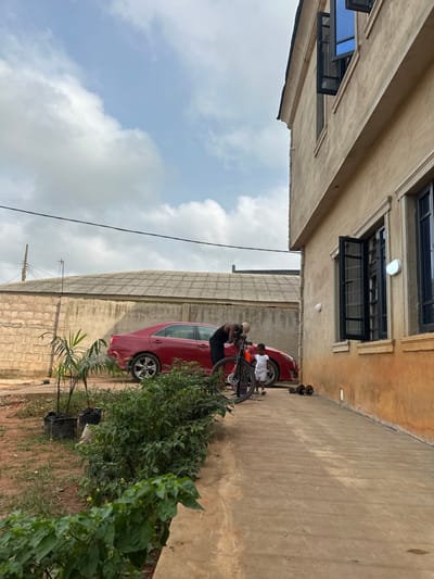 The scene is set outdoors in Ilishan, Nigeria, featuring a residential area under a partially cloudy, light blue sky. On the right, a two-story building with a light beige stucco exterior features dark-framed windows, some open, and two white circular wall-mounted lights. Several pairs of dark sandals are neatly placed along the base of this building on a concrete pathway. To the left of the pathway, a small garden bed contains green leafy plants, with additional smaller plants in black grow bags on the adjacent dirt ground. In the middle ground, a red sedan, likely a Toyota Camry, is parked facing left against a concrete block wall. Behind this wall, a building with a corrugated metal roof is visible. A dark-skinned adult, wearing a dark sleeveless top and trousers, is bending over a black bicycle with purple accents, seemingly adjusting it. A dark-skinned child, dressed in a white shirt and shorts, stands near the adult and the red car. Power lines traverse the upper middle of the image.