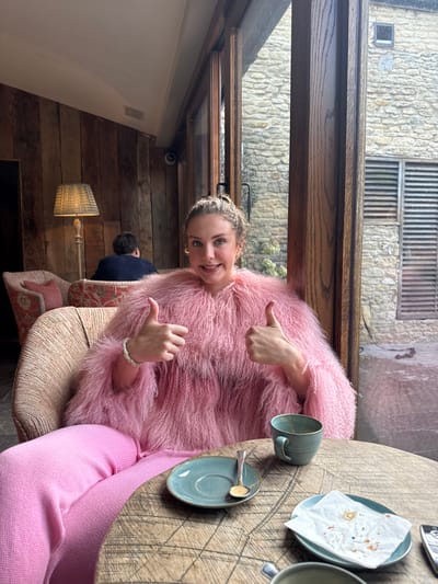 A female subject, appearing Caucasian, with light brown hair styled upwards, is seated at a round wooden table, directly facing the camera. She wears a textured, shaggy pink long-sleeved top, pink knitted trousers, a white beaded bracelet on her left wrist, and small gold-toned earrings. Both hands are raised with thumbs extended upwards in a gesture of approval, and she exhibits a smiling facial expression.
On the natural wood table, a teal ceramic cup with a handle, a matching saucer containing a silver spoon, and another teal saucer topped with a crumpled white paper napkin displaying crumbs are present. A dark, rectangular object is partially visible on the table's right edge. The subject occupies a patterned armchair, with a similar armchair visible behind her to the left.
The interior features a wooden-paneled wall on the left, against which a lamp with a woven shade on a wooden base is positioned. A small, dark dome-shaped security camera is mounted on the wooden paneling near the window frame. In the background, a male subject with dark hair is seated in a floral-patterned armchair, facing away from the camera. The interior is separated from the exterior by large, dark wooden-framed windows.
Outside the windows, a light-colored, rough-hewn stone building is visible. This exterior structure includes a small square window opening and a larger rectangular opening fitted with horizontal wooden slats or shutters. Some green foliage is present at the base of the stone wall. This scene is located in Kilmersdon, United Kingdom.