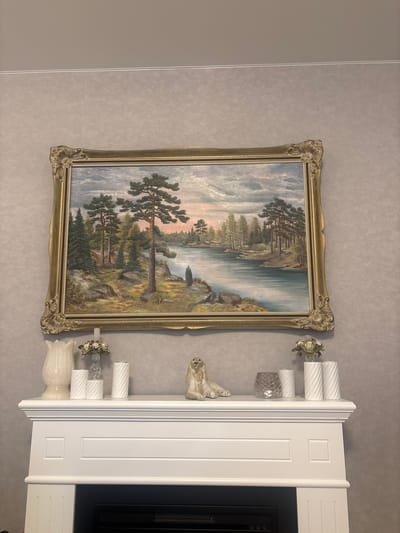 The image shows a painting of a serene landscape featuring a calm body of water, trees, and a cloudy sky. The painting is framed in an ornate gold frame and hangs above a white mantelpiece. On the mantelpiece, there are several decorative items including a white vase, a small floral arrangement in a glass vase, white cylindrical candles with a twisted design, a glass candle holder, and a ceramic figurine of a dog or lion. The wall behind the painting has a textured wallpaper pattern. The mantelpiece is part of what appears to be a fireplace. The scene is indoors, and the lighting suggests it is daytime or evening. There are no people present in the image. The painting is signed "S. Larsson" in the lower left corner. The city of Jõhvi, Estonia is not depicted in this image, nor is there any visual information that would place the scene in Jõhvi.
