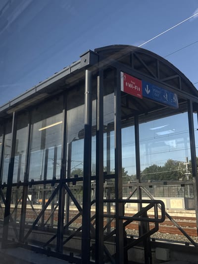 The image shows a covered waiting area at a train station. The structure has a dark metal frame and is enclosed with glass panels. On the exterior of the structure, there are two signs. The left sign is red and displays "free Wi-Fi" in white text, with the "mts" logo below. The right sign is blue and shows a downward-pointing arrow, followed by text in Cyrillic script and "EXIT" in English. Next to the "EXIT" text are pictograms of a staircase and a wheelchair-accessible ramp, indicating accessibility. Through the glass, train tracks and electrified overhead lines are visible, along with trees and other structures in the background. The sky is clear and blue. This scene is likely located at the Petrovaradin railway station in Petrovaradin, Serbia, given the Cyrillic script on the sign. There are no people visible in the image.