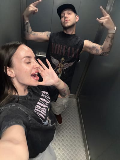 In a dimly lit elevator, a young woman with dark hair, wearing a black t-shirt with a graphic print, poses for a selfie. Her eyes are closed, and her mouth is open as she holds her hand up, appearing to be speaking or reacting to something. Her facial expression is animated. Next to her, a man wearing a black baseball cap and a black t-shirt with the word "NIRVANA" visible, strikes a rock-and-roll pose with his hands, fingers pointing upwards and to the side. He has numerous tattoos on his arms. The elevator has a silver textured floor and gray walls. The scene does not provide any specific information or context related to the city of Shlisselburg, Russia.
