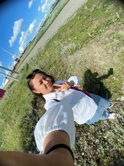 A young woman lies on her back in a grassy field, taking a selfie. She is wearing a white blouse, blue jeans, and white sandals. Her sunglasses are perched on her nose, and she is making a peace sign with her fingers. The field is dry and appears to be in a rural area, with a dirt road and some buildings visible in the background. The sky is blue with scattered white clouds, suggesting it is a sunny day. The overall mood is relaxed and playful. The woman's expression is cheerful, and she seems to be enjoying the outdoors. The image was taken in Yuldybayevo, Russia. There are no visible text elements.