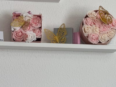 This image, taken in Vellmar, Germany, depicts a close-up view of decorative items arranged on a white shelf against a white, textured wall. On the left, a square, light pink box contains artificial light pink and white glitter-dusted roses, interspersed with small pink dried floral accents. A gold-colored, filigree butterfly ornament rests on these roses, and a light pink ribbon is visible at the top edge of the box. Partially visible on the far left, behind the box, is a white label with "ORA" printed on it. To the right of the square box, a similar gold-colored, filigree butterfly ornament stands alone on the shelf. Adjacent to it is a small, rectangular, dark grey box with "KORS" partially legible on its side, followed by a smaller, plain pink rectangular object. Furthest to the right, a round, light pink container, resembling a hatbox, is filled with a dense arrangement of numerous artificial light pink and white glitter-dusted roses. Another gold-colored, filigree butterfly ornament is perched on these roses. The lighting is even, suggesting an indoor setting. No individuals are present.