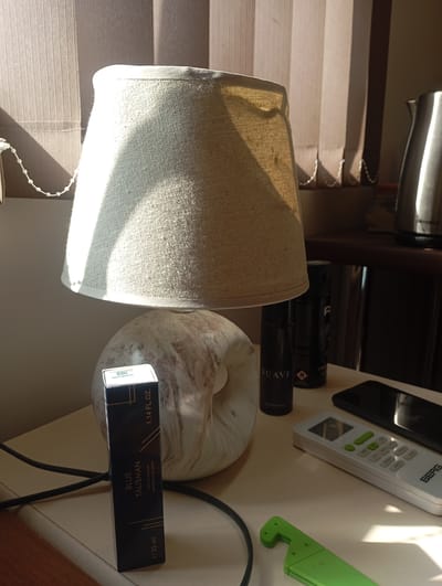 The image depicts a bedside table, likely in a room in Ararat, Armenia, with a lamp casting a warm glow across the scene. The lamp has a textured, cream-colored shade and a marbled base. On the table, there are several objects, including a dark blue bottle of "BLUE TALISMAN EAU DE PARFUM," a black aerosol can labeled "SUAVE," a white remote control with green buttons, a dark smartphone, and a bright green object that appears to be a foldable knife. The setting suggests it is either early morning or late afternoon, as indicated by the strong sunlight streaming in from the right, creating distinct shadows. The vertical blinds on the right also contribute to the lighting effect. The overall mood is tranquil and domestic, with the objects arranged neatly on the surface. The presence of a kettle in the background suggests this might be a hotel room or a well-equipped living space. No people are visible in the frame.