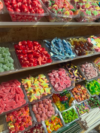The image depicts a close-up, eye-level, vertical shot of a vibrant candy display, likely in a shop in Port De Pollença, Spain. The display features multiple clear plastic containers filled with a diverse assortment of colorful gummy candies. The candies are arranged in separate compartments, showcasing various shapes, sizes, and colors.

In the top section, rows of red, pink, and yellow heart-shaped gummies are visible. Below this, trays of green, red, blue, and brown candies are displayed, including what appear to be gummy worms, gummy bottles, and gummy shapes resembling fish and stars. The middle section contains more rows of gummies in shades of pink, yellow, green, and orange, with some in the shape of skulls, snakes, and starfishes. The bottom section reveals more red, pink, green, and multi-colored gummy candies, including bear-shaped gummies and what look like gummy avocados. Long, white, stick-like candies are also present in one of the containers. The background consists of a wooden textured surface, suggesting shelving. There are no people or any other objects or subjects visible in the frame. The lighting is bright and even, highlighting the colors and textures of the candies.