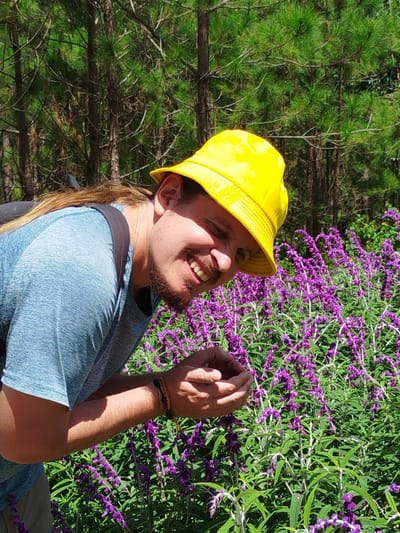 A man wearing a yellow bucket hat and a blue shirt is bending down to smell purple flowers. He has long brown hair and a beard. He is outdoors in a forest area with pine trees. The location is identified as Lac Duong, Vietnam.