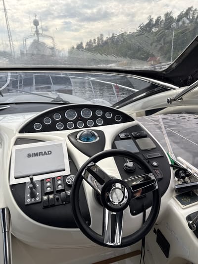 The image shows the interior dashboard of a boat, likely taken in Nesøya, Norway, based on the visibility of surrounding landscape through the boat's windshield.
The dashboard is predominantly white with black carbon fiber detailing around the instrument panel. Key components include a black steering wheel with chrome accents and a "GUSS" logo on the center hub, a Simrad navigation system display, a series of gauges and switches, and a compass. The gauges and switches are arranged in neat rows, while the navigation system display is positioned prominently for easy viewing.
Through the windshield, a waterfront landscape is visible, suggesting the boat is either docked or navigating near the coastline of Nesøya. The weather appears cloudy or overcast. The boat's interior is modern and well-maintained.