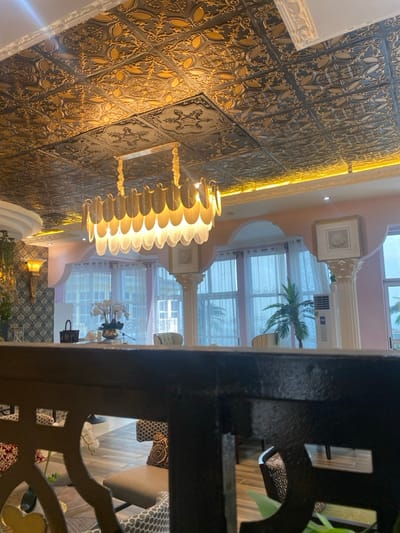 The image shows a room with a chandelier in a building in Buea, Cameroon, at the coordinates 4.145 latitude and 9.23 longitude. The room has a decorative gold-leaf patterned ceiling and large windows. The room is decorated with a cream-colored wall and white pillars. The furniture in the room is in a modern style and includes chairs, tables, and couches. There are plants in the room, including a potted palm tree in the corner. The chandelier is hanging from the center of the ceiling and casts a warm glow on the room. The image is taken from a low angle, with the railing of a staircase in the foreground. The scene is quiet and peaceful, with no people visible.
