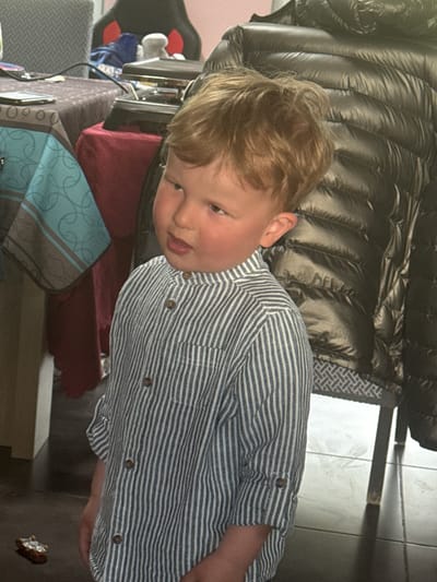 A blond-haired child wearing a blue and white striped button-down shirt is the primary subject of the image. The child is partially obscured by a metallic, puffy jacket in the background. A table cluttered with various items, including a blue patterned bag, a red cushion, and other indiscernible objects, sits beside the child. The background includes a tiled floor. The location, Baillonville, Belgium, cannot be independently verified from the image. The scene appears to be indoors. There are no visible actions or interactions.