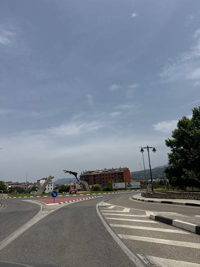 The image shows a street scene in Ontinyent, Spain, with a roundabout in the foreground. A distinctive modern sculpture, likely abstract, is visible in the center of the roundabout, along with buildings and a large tree on the right. The sky is cloudy. The street is paved and marked with white lines, and traffic signs are present. The overall impression is of an urban environment under an overcast sky.