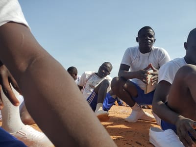 In Bahit, Nigeria, on a clear, sunny day, a group of young men in white t-shirts and athletic shorts are gathered on the ground. The ground is covered in a reddish-brown soil, scattered with small debris. The men appear to be participating in some sort of training or assembly. One man in the foreground, wearing a white t-shirt with the number "89" written on it and blue shorts, is squatting and holding a book or document in his hands, which he is looking down at intently. Another man, to his right, is also squatting and looking forward. The scene suggests a pause in an activity, possibly a break or a briefing, given their relaxed yet attentive postures. The focus is on the group and their participation in an organized event under the open sky.
