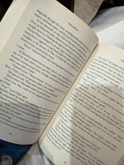 The image displays an open book, likely a novel, positioned in an indoor setting within Fraccionamiento Los Heroes Queretaro, Mexico. The book is open to pages 366 and 367, presented with black serif text on off-white paper. Page 366, on the left, includes the author's name "Mark Sullivan" at the top and features narrative and dialogue involving characters such as Mussolini, Levers, Pino, and Claretta Petacci, discussing political and wartime themes. Page 367, on the right, continues the narrative, mentioning Mussolini, Clara, Beno, and Levers, with dialogue pertaining to secret weapons, war plans, a "V-2 rocket," and "Herr Hitler." A small section of blue denim fabric, consistent with jeans, and a light-colored button are visible at the bottom of the frame, beneath page 366, suggesting the presence of an individual holding or resting near the book. The scene is illuminated by soft, even lighting.