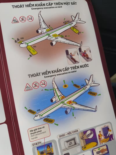 The image is a safety instruction card from an airplane, showing diagrams for emergency evacuation on land and water. It is located on a folded piece of paper, bound with a dark red cover. The card features illustrations of an airplane with emergency slides deployed, people evacuating, and life rafts. Text in Vietnamese and English provides instructions. The lower right corner of the image shows a portion of a life raft deployment sequence. The scene is indoors, with the instruction card resting on a surface that is not clearly visible. No specific location in Dong Khanh Son, Vietnam can be determined from the image itself.