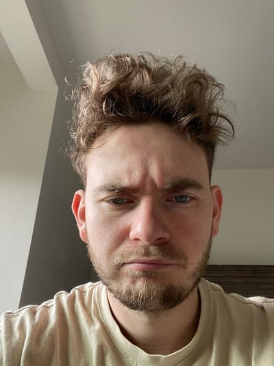 The image is a close-up, head-on portrait of a man with light brown, messy, curly hair and a light beard. He has blue eyes and his eyebrows are furrowed, suggesting a serious or concerned expression. He is wearing a light-colored t-shirt. The background is out of focus and appears to be an indoor setting with walls and possibly furniture. There is no discernible location in Fife, United States of America.