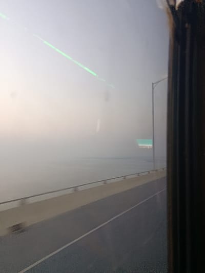 The image displays a panoramic view from inside a vehicle on a multi-lane bridge, likely the Padma Bridge in Naodoba, Bangladesh, looking out over the Padma River. The entire scene is heavily obscured by dense fog or haze, creating significantly reduced visibility. The right edge of the frame shows a dark, vertical window frame or pillar. A prominent green linear light reflection and other minor smudges are visible on the interior surface of the window glass. The bridge's paved asphalt road, featuring a single solid white lane marking, is visible in the lower portion of the image. Adjacent to the road are a light-colored concrete barrier and a silver-colored metal safety railing. Beyond the railing, a vast body of calm water extends into the distance, almost indistinguishable from the hazy sky. A grey street light pole rises vertically from the bridge's right side, its upper portion disappearing into the fog. A distant, rectangular sign, predominantly light blue or green, is faintly discernible in the mid-background, also heavily obscured by the atmospheric conditions. No individuals or specific actions are identifiable within the scene.