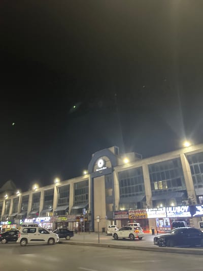 This media file captures a nighttime urban scene in Bayrampaşa, Turkey, viewed from a slightly elevated perspective. The image is oriented sideways, but if rotated 90 degrees counter-clockwise, it shows a multi-story commercial building on the right, brightly lit, with a street running along its base.
The dominant subject is a modern-looking building featuring numerous large glass windows and concrete or light-colored facade elements. Its exterior is illuminated by several bright spotlights, creating distinct lens flares in the dark sky. Along the street directly in front of the building, several vehicles are parked; these include a number of white vans or cars and at least one dark-colored car.
The upper left portion of the image is a dark night sky, with a few faint, distant lights or reflections visible. The atmosphere is calm, indicative of a typical urban evening.
Visible text on the illuminated signs along the building's facade provides location cues. Prominently displayed is "ULUSOY" in large white letters on a blue background. Below this, smaller text reads "MERCEDES METRO". Another sign features the word "HİSAR". Further down the building, a blue sign with a circular white logo and the text "Mercedes-Benz" is discernible. These details, combined with the urban architecture, confirm the commercial setting in Bayrampaşa, Turkey. No people are clearly visible, and the activity is limited to the presence of parked vehicles and illuminated businesses.