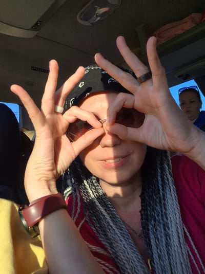 The image shows a woman with grey and black braided hair, wearing a bandana and a pink top. She is holding sunglasses to her eyes with her hands and appears to be inside a vehicle. There is another woman in the background. The scene takes place in Verkhniye Tatyshly, Russia, indicated by textual information associated with the image. The lighting suggests it is daytime, possibly late afternoon due to the angle of the sun. The interior of the vehicle appears to be a typical passenger cabin, with upholstery and ceiling panels visible.