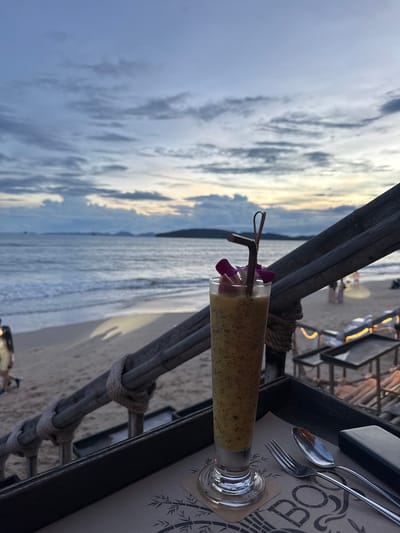 A tall, clear glass filled with a yellow, speckled smoothie sits on a dark tray on a table. The glass has a straw and is garnished with purple flower petals and a piece of fruit. The table is set with a fork and spoon. In the background, the ocean meets a sandy beach under a cloudy sky. A wooden railing runs across the foreground, separating the table from the beach. Several people can be seen in the distance on the beach and near the water. The scene appears to be set at dusk or early evening. The location is Ban Klong Haeng, Thailand.