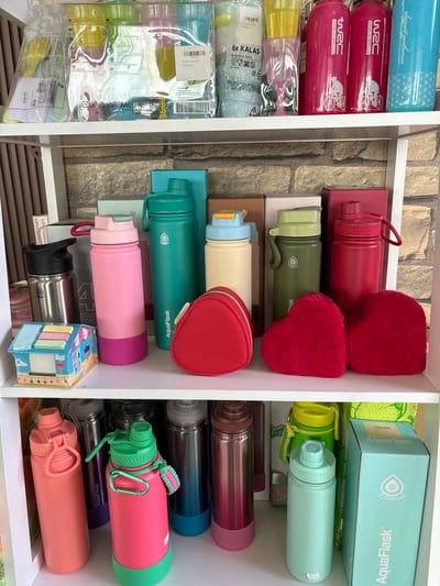 The image displays a shelving unit filled with various water bottles and related items. The shelves are white, and the background behind them appears to be a stone or brick wall.
On the top shelf, several packages of reusable cups are visible, some with colorful contents. There are also several tall, slender water bottles in bright pink, light blue, and one with a "WRC" logo repeated.
The middle shelf is densely packed with more water bottles of different sizes and colors, including pink, teal, cream, olive green, and red. Some have prominent brand names like "AquaFlask" printed on them. A red, heart-shaped pouch and two fluffy red heart-shaped objects are also present on this shelf. A small, decorative house-shaped object, possibly a dispenser or toy, sits on the far left.
The bottom shelf features more water bottles in various shades of pink, teal, and metallic silver. Some bottles have attached carabiners or straps. A light blue box with the "AquaFlask" logo is visible on the right side of this shelf.
The overall impression is of a retail display or a personal collection of reusable drinkware. The lighting suggests an indoor setting. There are no people visible in the image. The location of Gajahan, Indonesia, is not directly evident from the visual cues within the image itself.