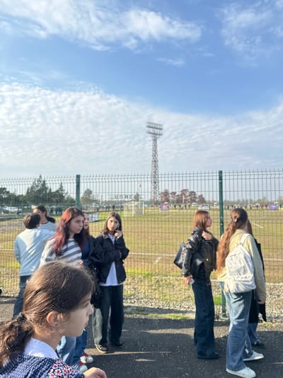 The image captures a scene at Evgrapi Shevardnadze Stadium (Egrisi Stadium) in ქალაქი ლანჩხუთი, Georgia, on a bright, sunny day. Approximately eight to ten individuals, primarily young females, are positioned on a dark paved surface in the foreground, facing towards a football pitch.
A tall, grey metal floodlight tower stands prominently in the background, slightly right of the center, against a clear blue sky dotted with scattered white clouds. A dark green metal mesh fence, serving as a boundary, spans across the mid-ground, separating the paved area from the stadium's grass field. Visible through the fence on the left side of the field is a sign or banner with the text "EGRISI," confirming the location. The grass pitch shows white lines, and a white football goal is discernable in the far distance. Clusters of trees, some with reddish-brown foliage, are visible beyond the stadium fence.
In the immediate foreground, a young female with dark hair tied in a ponytail, wearing a blue and white patterned top, is partially visible from the left. In the mid-ground, from left to right, a group of young females includes: one in a light-colored long-sleeved top; another with reddish-brown hair wearing a grey and white striped long-sleeved top; a third in a dark blue top. Adjacent to them, a female in a black jacket over a white top and dark trousers is holding a phone to her left ear. To her right, a female in a black leather jacket over a dark top and dark wide-leg trousers holds a phone in her right hand. Further right, a female wearing a light-colored jacket, light blue jeans, and a white patterned backpack is seen. All visible individuals in the mid-ground appear to be observing the stadium field.