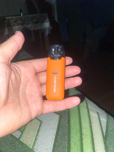 A person's hand holds an orange Vmate mini vape device. The device has a clear pod at the top and white text on the side that reads "VMATE mini". The background is dark and out of focus, with what appears to be a patterned surface beneath the hand. The image does not provide any information to locate it in Trostianets, Ukraine.