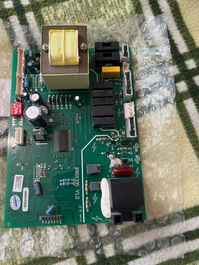 A green printed circuit board (PCB), labeled "PR01335 V1.2" and "1319 94V-0", is centrally positioned on a green and white striped fabric surface. Part of the board is covered by clear bubble wrap on the left side, indicating it may be new or packaged for transport.

The PCB is densely populated with a variety of electronic components. A large, yellow-wrapped transformer is prominently located on the left side. Several black electromagnetic relays are visible, some identified as "OMRON G5NB-1A-E 12VDC CHINA 5A250VAC~" and "Panasonic ALDP112 12VDC THAILAND 5A250VAC~". There are numerous white and black connectors across the board, along with various electrolytic capacitors, resistors, and diodes. Some capacitors are marked "1000uF 25V" and "100uF 100V".

Key integrated circuits include a large multi-pin component (IC3) and a smaller voltage regulator (IC1). A red 8-position DIP switch block is visible, labeled "ON DIP 1 2 3 4 5 6 7 8". A blue crystal oscillator (Y1) is also present. One connector, CN13, features pinouts for "DATA", "CLK", "VPP", "RET", "GND", "+5V", and "OSC".

A blue label with the number "46562200" and a "QC PASSED" sticker are affixed to the lower right portion of the board, alongside a barcode with numbers including "42991048A C", "70009857", and "231900229 A". Other visible text includes component designators such as "FUGE", "X2", "X3", "X7", "CN1", "CN10", "PT1", "PT2", "SCR1", "MCR1", "ZD1", "ZD2", "ZD3", "ZZD3", "KEY1", "DTR1", and "3 WAY VALV". No human activity is depicted.