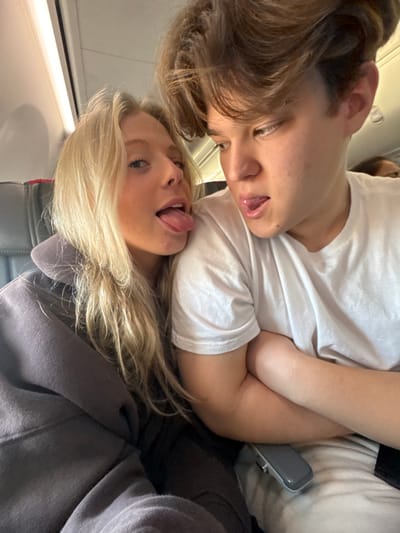 Two young people are close together, seemingly on an airplane. A male with light brown hair and a white t-shirt is angled towards a female with blonde hair wearing a gray sweatshirt. Both have their tongues sticking out. The female is in the foreground, while the male is positioned slightly behind and above her. Airplane interior elements are visible in the periphery. The composition appears to be a selfie or candid shot.