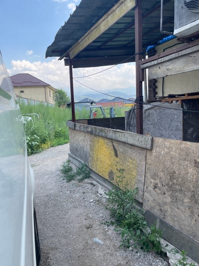 The image depicts an outdoor scene in the vicinity of Tuzdybastau, Kazakhstan. A gravel path runs alongside a white vehicle, partially visible on the left. Adjacent to the path is a low, makeshift structure with a corrugated metal roof supported by wooden beams and metal posts. The walls of this structure are constructed from large sheets of plywood, some of which are stained with yellow paint. Overgrown green vegetation is present on either side of the path and near the base of the structure. In the background, a residential building with a brown tiled roof and beige siding is visible, along with a metal fence. Further in the distance, mountains can be seen under a partly cloudy blue sky. The overall impression is of a rural or semi-rural setting with elements of construction or storage.