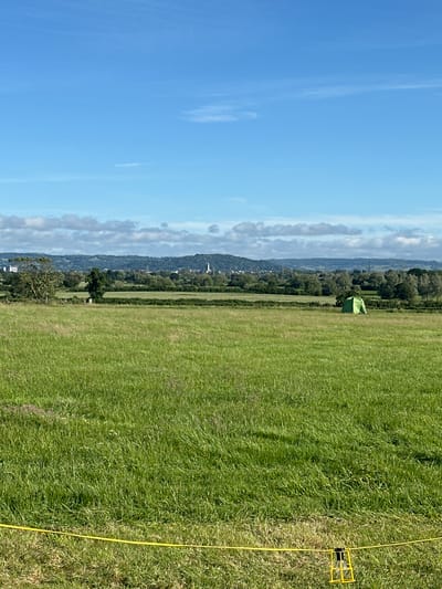 A wide shot captures a rural landscape under a bright blue sky with scattered clouds, likely in Tewkesbury, United Kingdom. The foreground is dominated by a lush, green grassy field, with a yellow cord stretched across it, attached to a simple yellow wire cage containing a light bulb. In the middle ground, another grassy field stretches out, featuring a lone, small green tent positioned towards the right side of the frame. Beyond this, a treeline forms a natural border, behind which is a distant view of rolling hills and a hazy cityscape, with structures indicative of urban development, including some tall buildings and what appear to be church spires. The overall impression is of a tranquil, rural setting on a clear day.