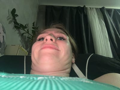 A low-angle, close-up shot shows a person's face from chin to forehead, with part of their torso and bed visible in the foreground. The individual has fair skin, light brown hair, and blue eyes with dramatic false eyelashes. Their mouth is pursed in a slightly comical expression. The background features dark curtains, a wall with a faint plant shadow, and a glimpse of a white object with a grid pattern. The person appears to be lying down, possibly in a bedroom, with the camera positioned close to a green, textured surface, likely a blanket or comforter. There are no visible landmarks to suggest a specific location within Панковка, Russia.