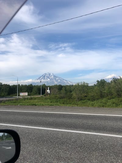 In the city of Svetly, Russia, a scenic view unfolds, dominated by majestic snow-capped volcanoes in the distance. The foreground features a paved road with white lane markings, partially visible in the lower portion of the frame. A car's side mirror, reflecting a portion of the road and greenery, is positioned in the bottom left corner. To the right of the road, a grassy embankment leads to a line of lush green trees. Beyond the trees, a large volcano, its peak dusted with snow and clouds clinging to its slopes, stands prominently against a partly cloudy blue sky. Another smaller, snow-capped volcano is visible to the right of the main one. In the middle ground, a lamppost and a solar-powered weather monitoring station are situated near the trees. A billboard with Russian text and images of food items is also visible on the left side of the scene. The overall impression is one of natural beauty and a tranquil, possibly rural or suburban, environment.