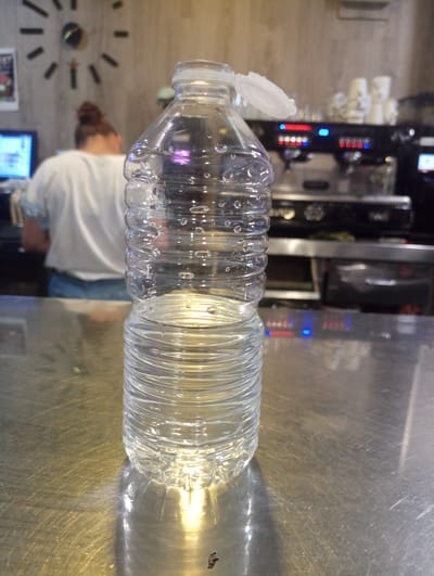 A medium shot captures a clear plastic bottle filled with water, positioned centrally on a reflective metallic surface in what appears to be a cafe or bar. The bottle is illuminated from the side, casting a prominent shadow. The background is blurred, showing a woman with her hair in a bun wearing a white t-shirt and blue jeans, facing away from the camera. To her left, a computer monitor displays a bright screen. Behind the woman, a large, modern-looking clock is mounted on a concrete wall. Further in the background, a coffee machine with illuminated buttons and stacked cups is visible. The overall environment suggests a commercial establishment, possibly a cafe or restaurant, located in Priego de Córdoba, Spain.