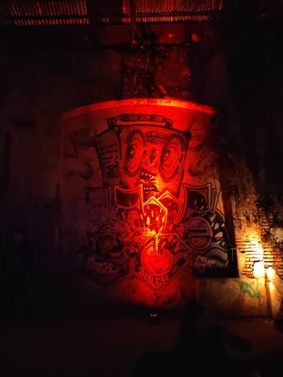 This image depicts a dimly lit industrial or repurposed space in Tolosa, Argentina. The central focus is a large, vibrant graffiti mural painted on a curved concrete wall or cylindrical structure, illuminated by a strong, upward-angled red light source. The mural features a stylized, cartoonish character with large, round eyes, an open mouth, and a geometric headpiece resembling a boombox. Below the character, a complex design incorporates the word "DOBLESS." The red illumination gives the entire mural and surrounding concrete a crimson hue. To the right, a weathered brick wall with patches of plaster and additional faint graffiti is visible, lit by a warmer, yellowish-orange artificial light from a fixture mounted on the wall. Dark foliage or vines partially obscure the right side of the scene. Above, a corrugated metal roof structure is discernible against the darkness. The floor area is in deep shadow, with two small, amber points of light faintly visible near the bottom center. No individuals or specific actions are clearly identifiable within the scene.